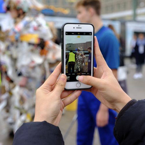Zwei Hände halten einSmartphone, das eine städtische Situation fokussiert. Die unscharfe Szene zeigt drei Menschen. Eine davon ist vermutlich mit Verpackungsmüll dekoriert.