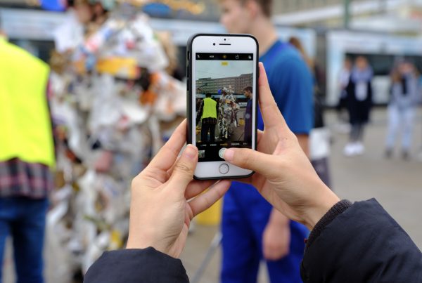 Zwei Hände halten einSmartphone, das eine städtische Situation fokussiert. Die unscharfe Szene zeigt drei Menschen. Eine davon ist vermutlich mit Verpackungsmüll dekoriert.