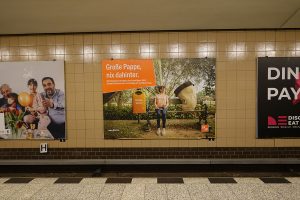 In einem Berliner U-Bahnhof hängt eine Plakatwerbung der BSR zur Stadtsauberkeit aus 2021. Darauf sitzt eine Frau mit einer Zeitung und einem Mehrwegbecher auf einer Parkbank. Ein überdimensional großer Papp-Einwegbecher schaut sie traurig an. Neben der Bank steht ein Papierkorb mit der Aufschrift „Becherwisser“. Die Headline der Kampagne lautet: „Große Pappe, nix dahinter.“ Die Subline löst auf: „Einwegverpackungen sind unnötiger Müll. Schone Ressourcen und steig um auf Mehrweg.“